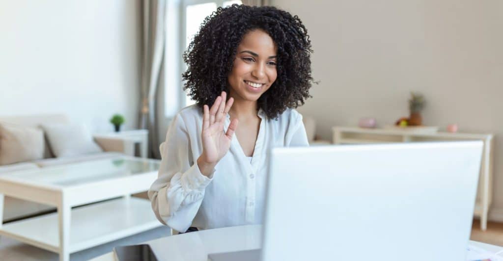 A woman with curly hair smiling and waving at a laptop screen during a video call, in a brightly lit room