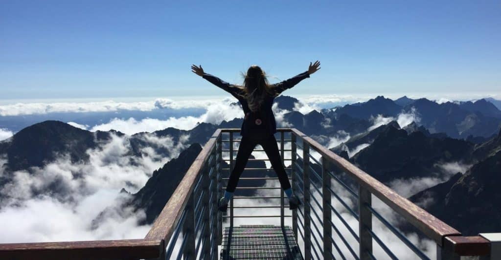 A woman with her arms outstretched, wearing a backpack, standing on a viewing platform overlooking a vast mountain range