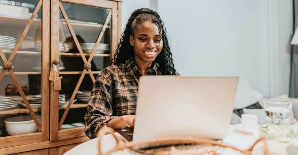 A smiling woman with braided hair using a laptop at a table in a home setting