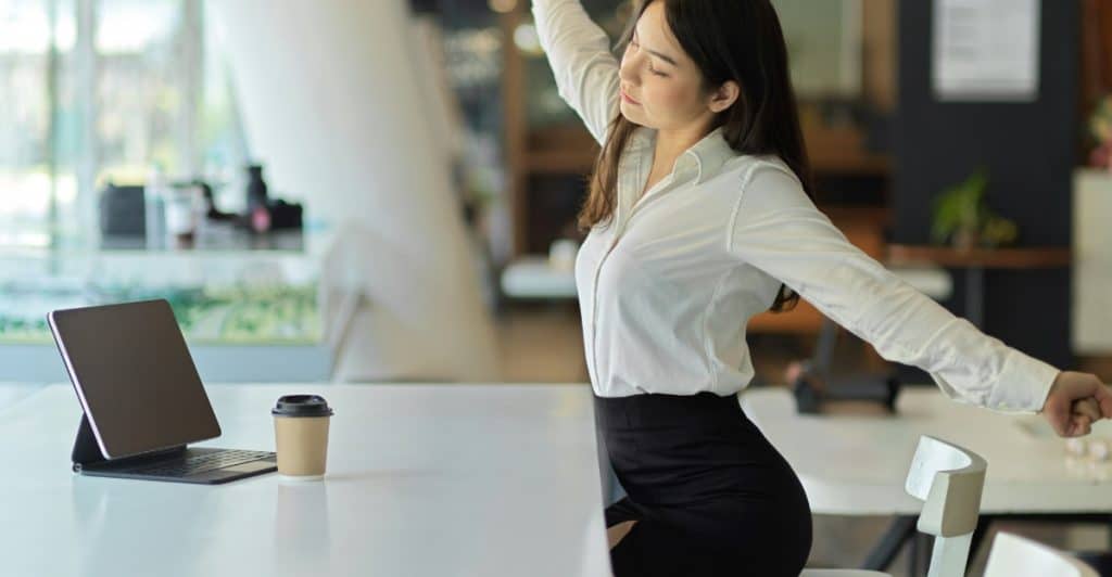 A woman in a white shirt and black skirt stretching her arms above her head while sitting at a desk with a laptop and coffee cup.