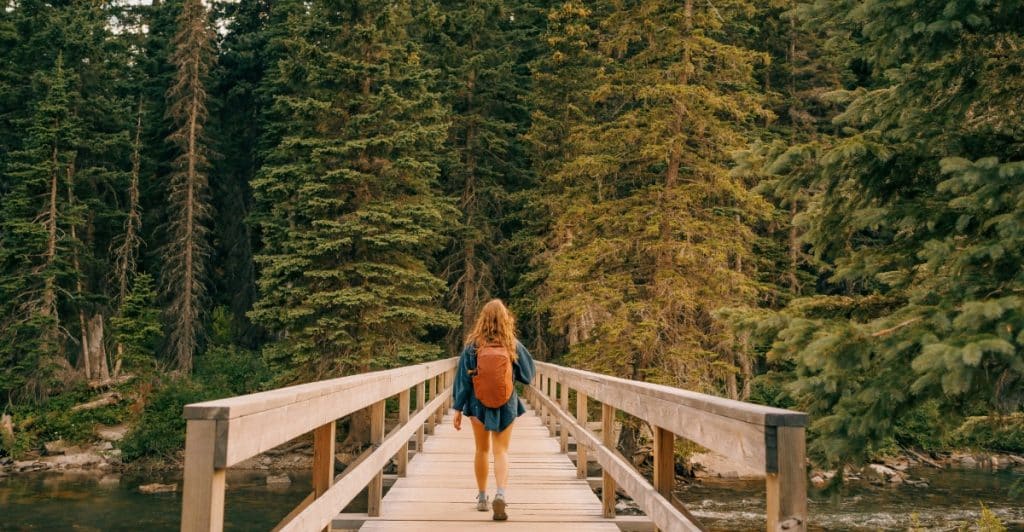 A woman with an orange backpack walking away on a wooden bridge through a dense forest.