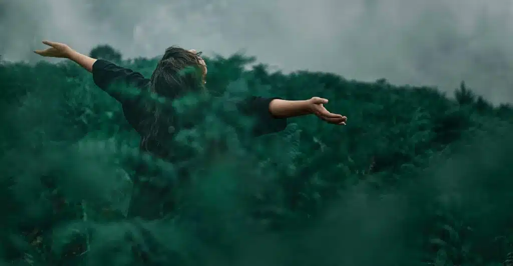 A woman from behind with long dark hair and a black top, standing with her arms outstretched amidst lush green foliage, embracing nature.