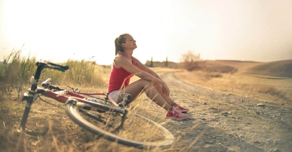 A woman sitting on the ground next to a bicycle in a rural outdoor setting, looking up with her eyes closed.