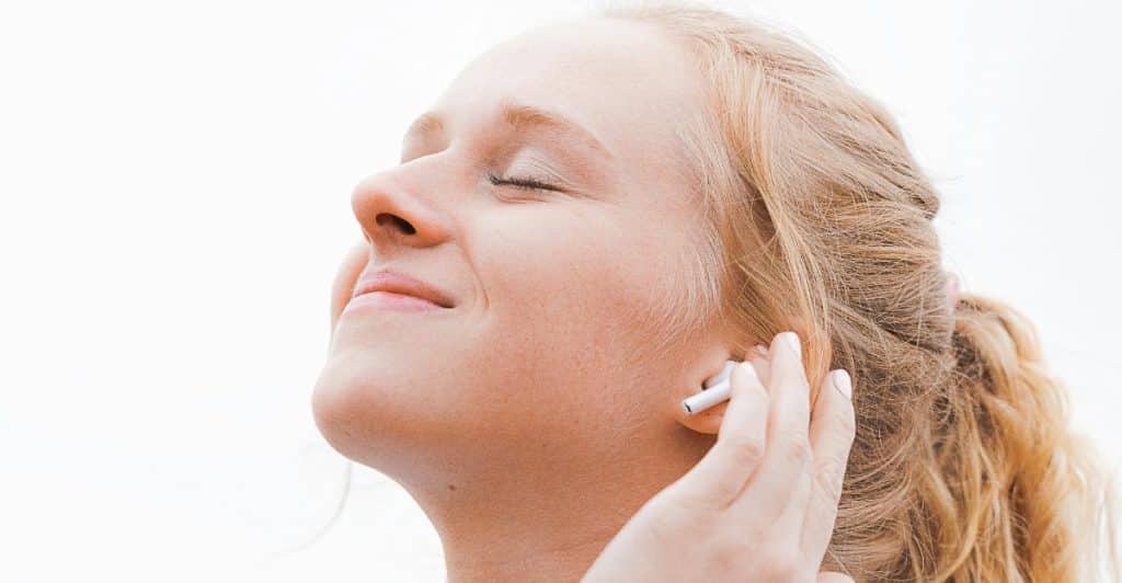 A woman with blonde hair and closed eyes, wearing white earbuds, touching her ear with one hand, against a light background.