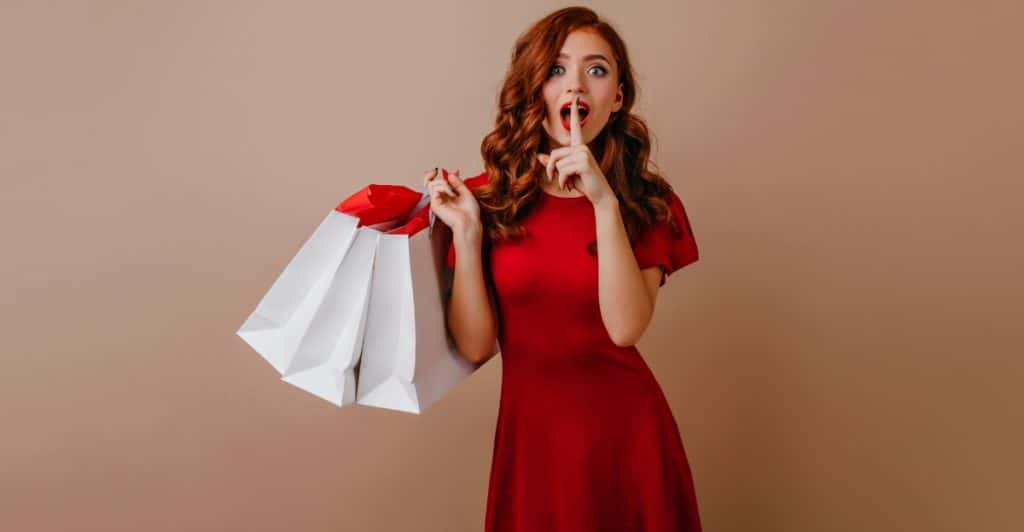 A woman with long red hair wearing a red dress, holding white shopping bags and making a shushing gesture with a surprised expression.