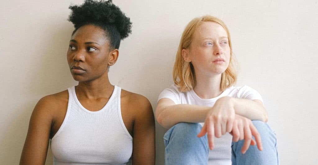 A woman with dark skin and dark hair sitting on the left looking away from a woman with light skin and blonde hair sitting on the right