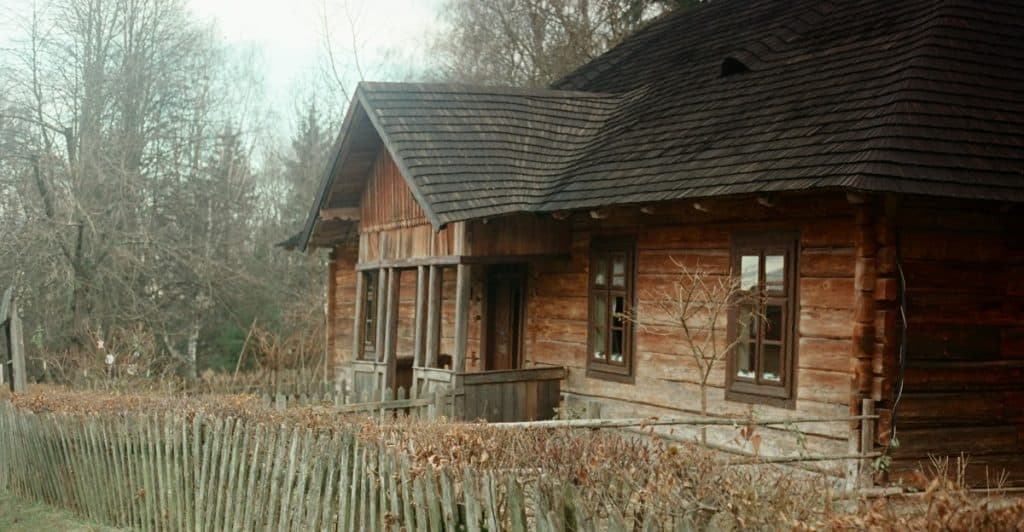A rustic wooden house with a fence surrounded by trees under a cloudy sky.