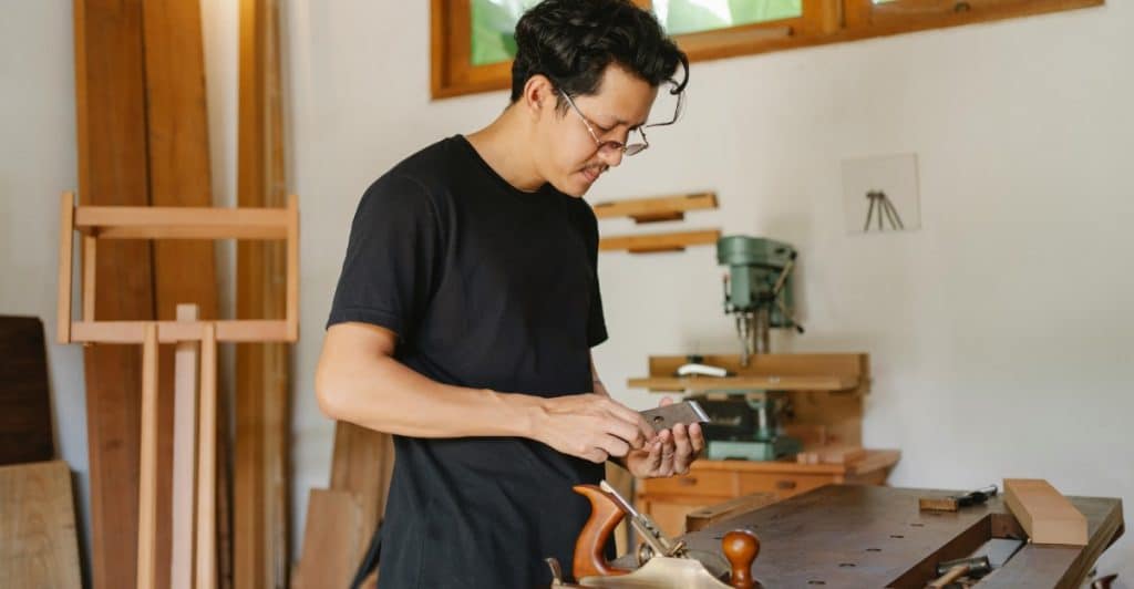 A man in glasses and a black t-shirt intently examining a small piece of wood in his hands, standing at a workbench with woodworking tools and unfinished wooden planks.