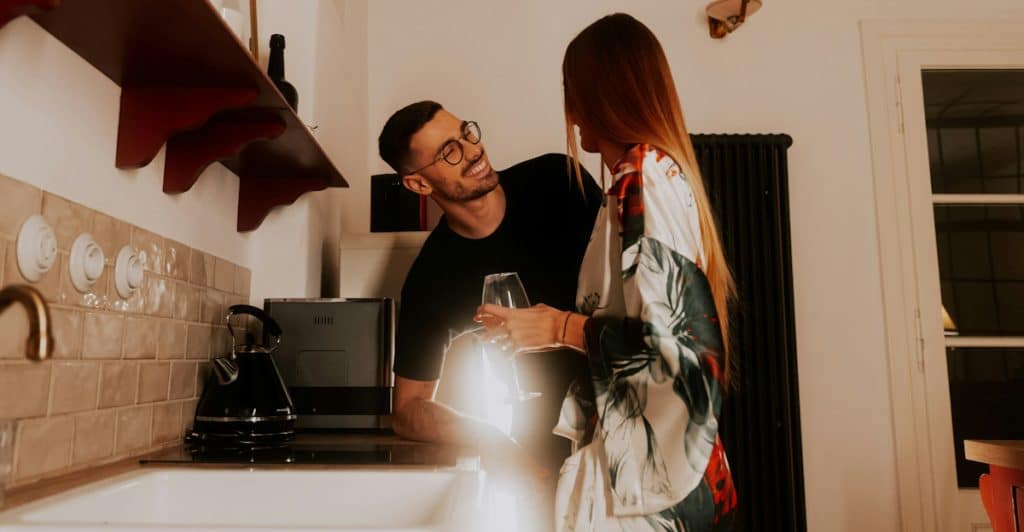A man with glasses leaning against a kitchen counter, smiling at a woman who is holding a wine glass in a cozy kitchen.