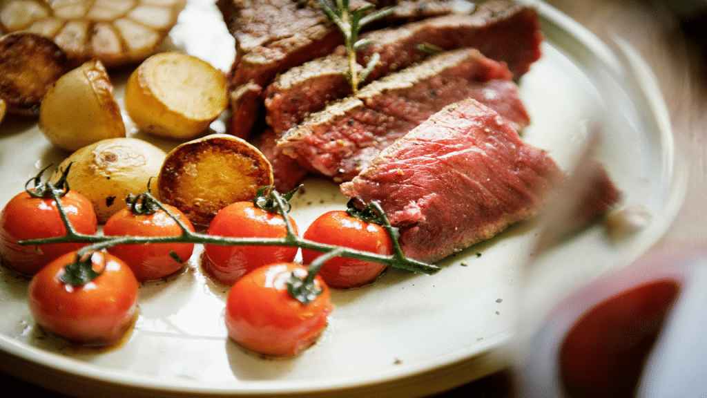 A plate of rare steak slices with roasted potatoes, cherry tomatoes, and garlic.
