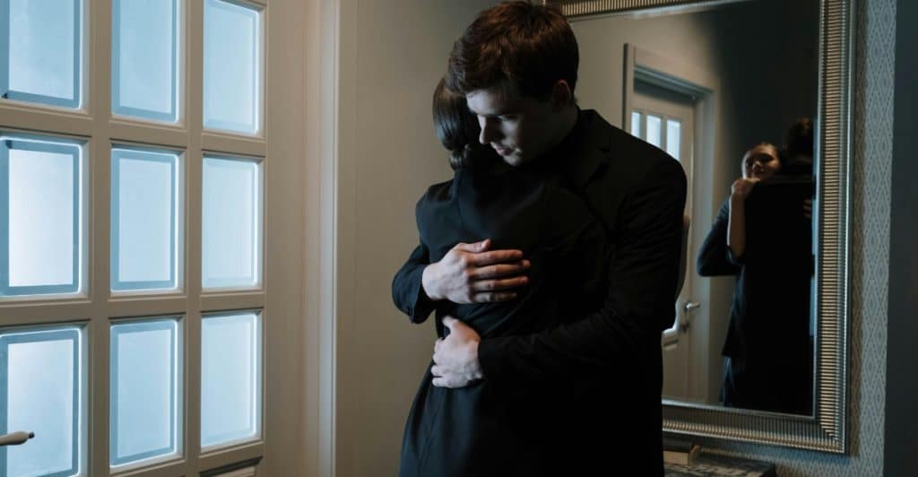 A man and woman dressed in dark clothing share an emotional embrace in a dimly lit hallway.