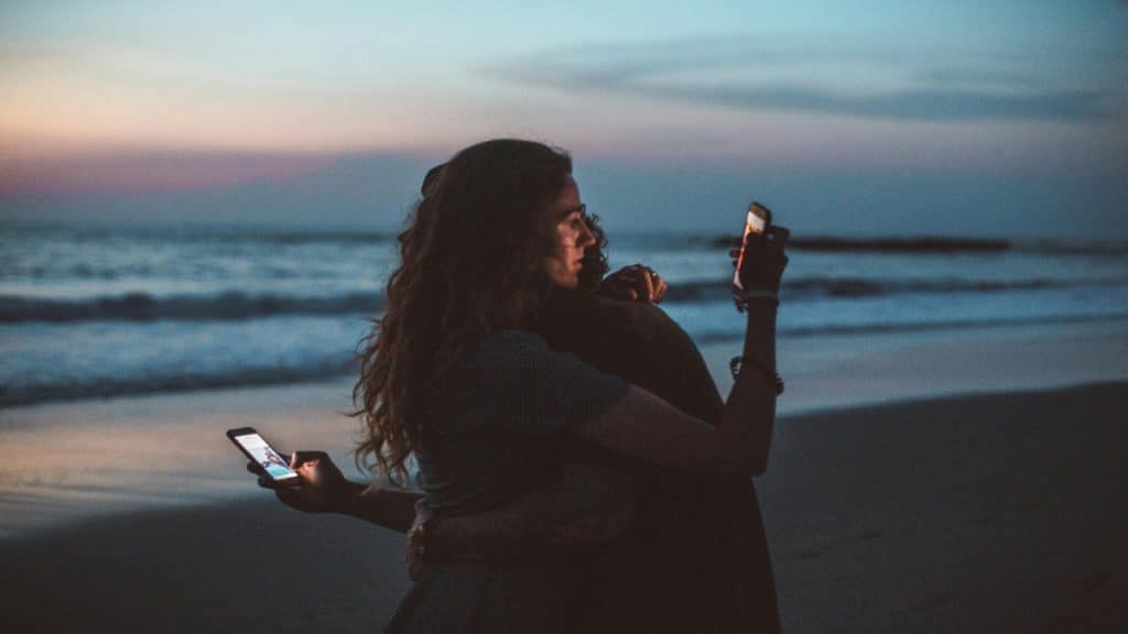 A couple hugging each other on the beach but absorbed in their phone.