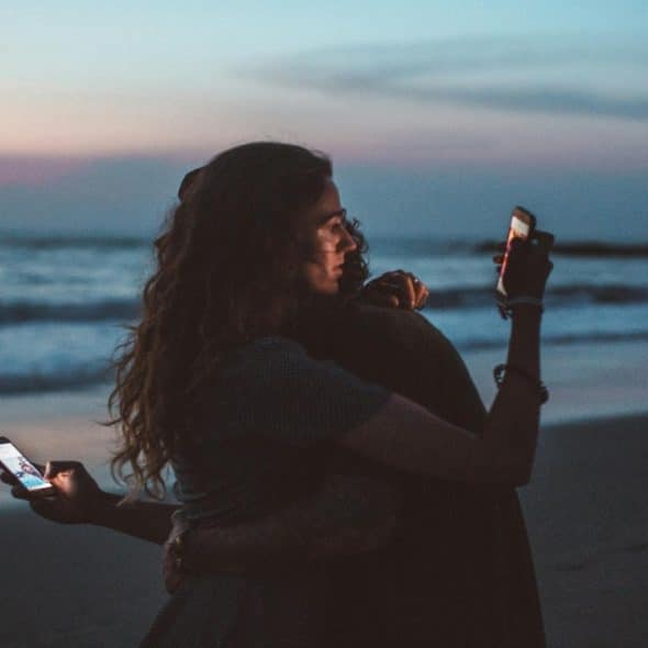 A couple hugging each other on the beach but absorbed in their phone.