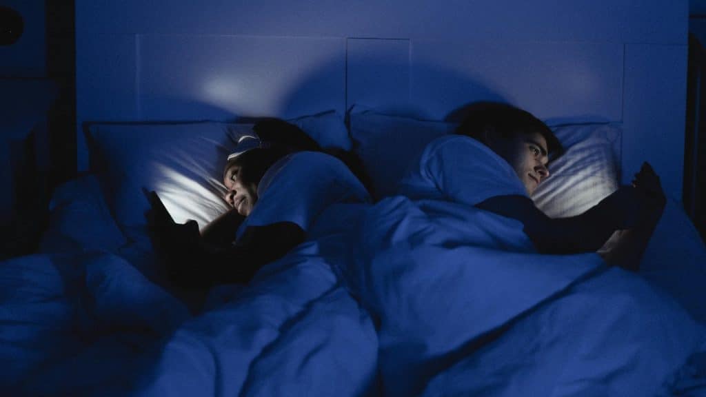 A couple lying back to back in bed using their phones, symbolizing overstimulation and lost intimacy.