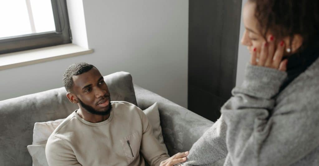 A man sitting on a gray couch gently holds the hand of a woman standing beside him.