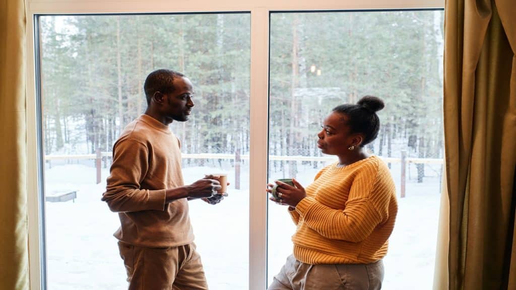 A couple stands near a window with a cup of coffee in their hand.
