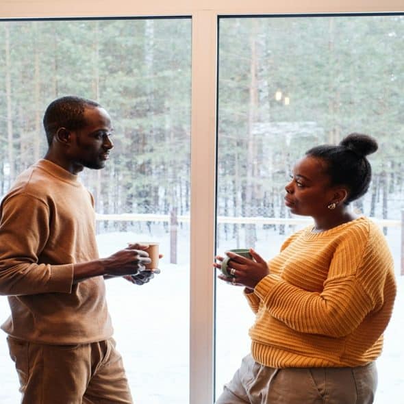 A couple stands near a window with a cup of coffee in their hand.