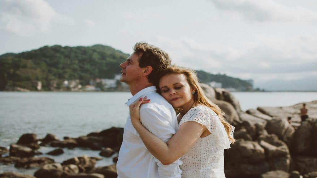 A woman hugs her partner from behind as they stand on the seaside rocks.