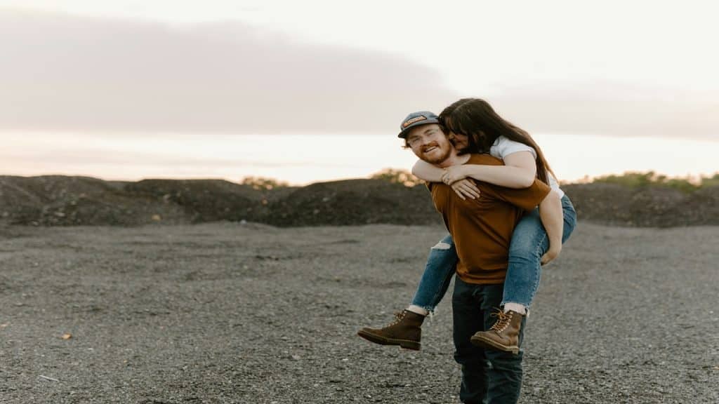 A woman jumps on her partner’s back as they walk on the sand.