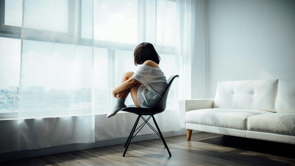 A woman sits curled up on a chair by a large window, gazing outside.