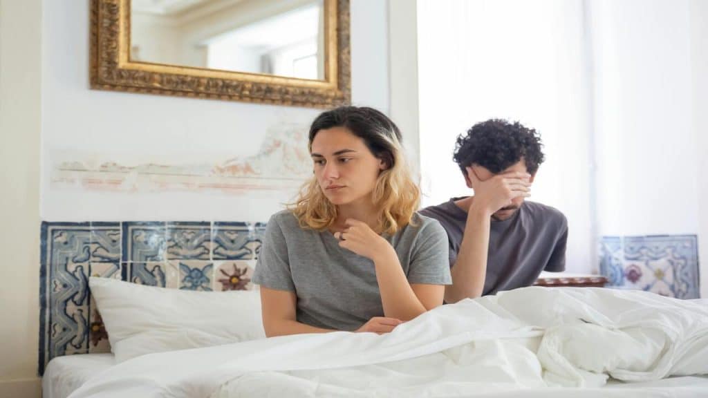 A woman and a man sitting on bed looking sad.