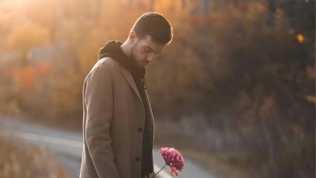 A man walking alone down a quiet street at dusk, holding a flower, deep in thought.