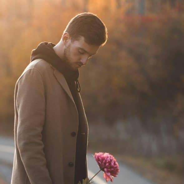 A man walking alone down a quiet street at dusk, holding a flower, deep in thought.