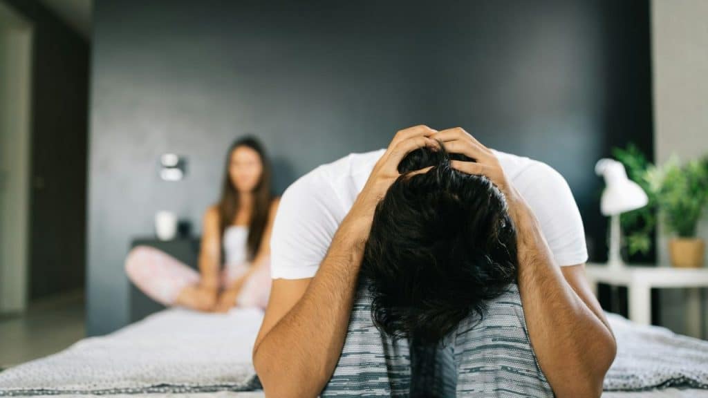 Man in foreground, head in hands, sitting on bed with woman in background.