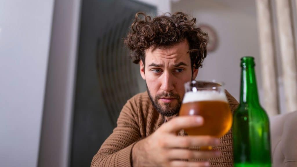 A man with curly hair looks at a glass of beer, a green bottle next to it.
