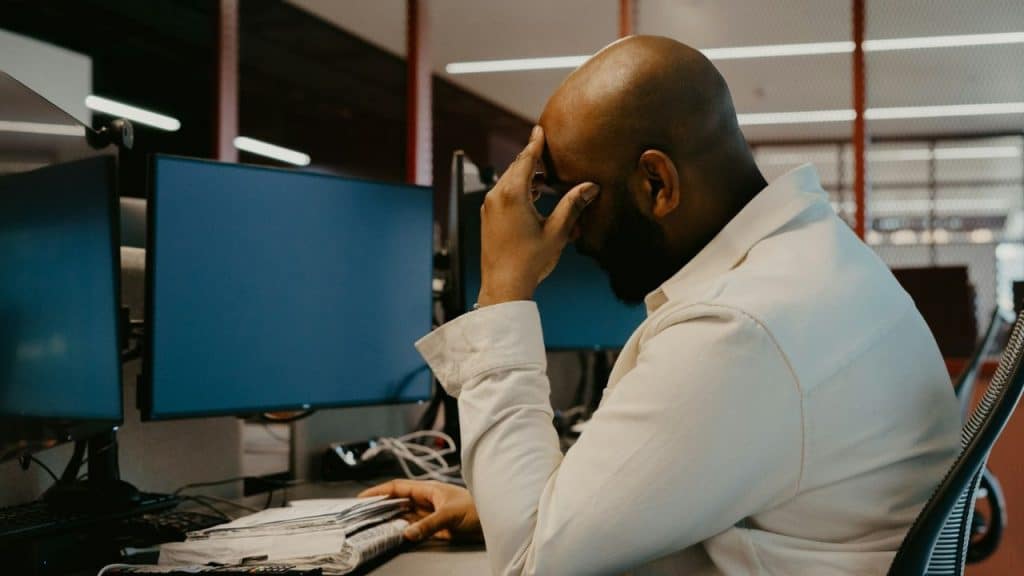A man sits at a desk with two monitors, holding his head in his hand.