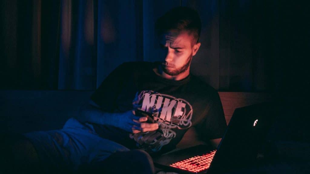 A man lies in a dark room, looking at his phone with a laptop nearby.