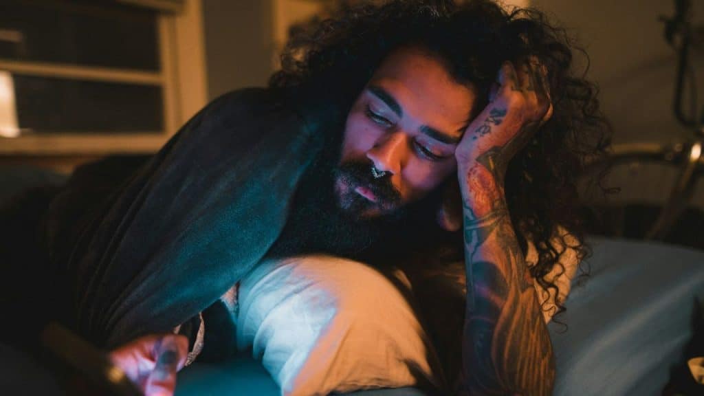 A person with long, curly hair and tattoos looks at a glowing phone in a dark room.