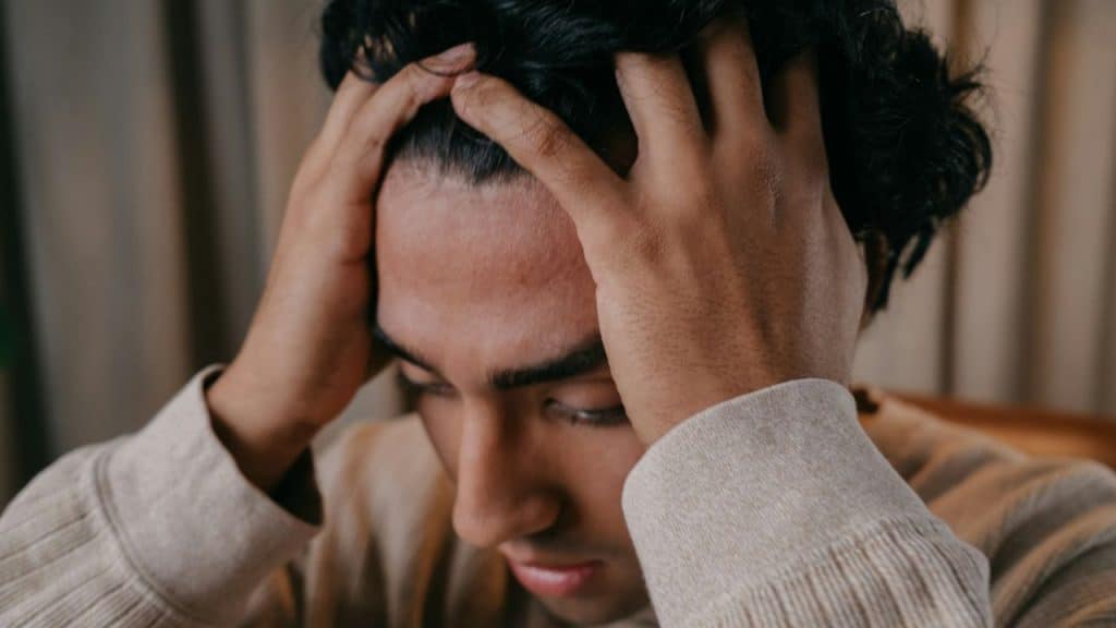 A man with dark, wavy hair holds his head with both hands, looking down.