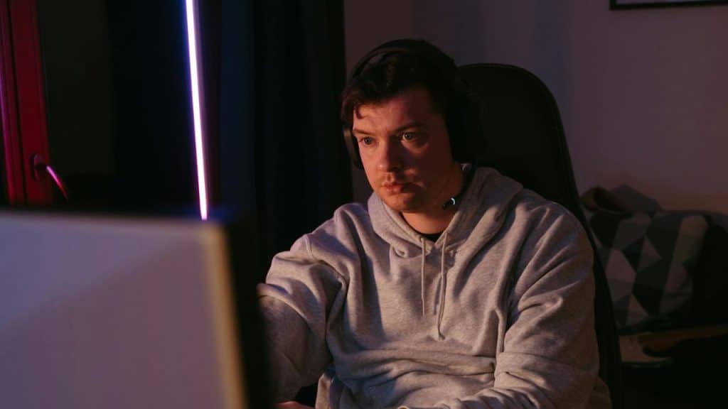 A man wearing headphones and a grey hoodie intently looks at a computer screen.