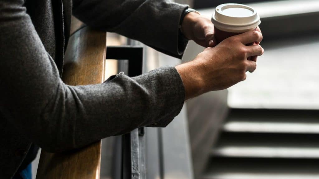 A man in a grey jacket holds a white-lidded coffee cup.