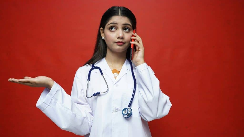 A female doctor in a white coat holds a phone to her ear, looking up and shrugging.