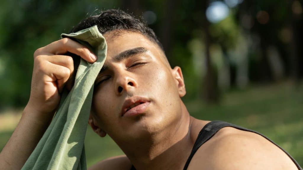 A person wipes their sweating forehead with a green cloth, eyes closed, in a sunny outdoor setting.