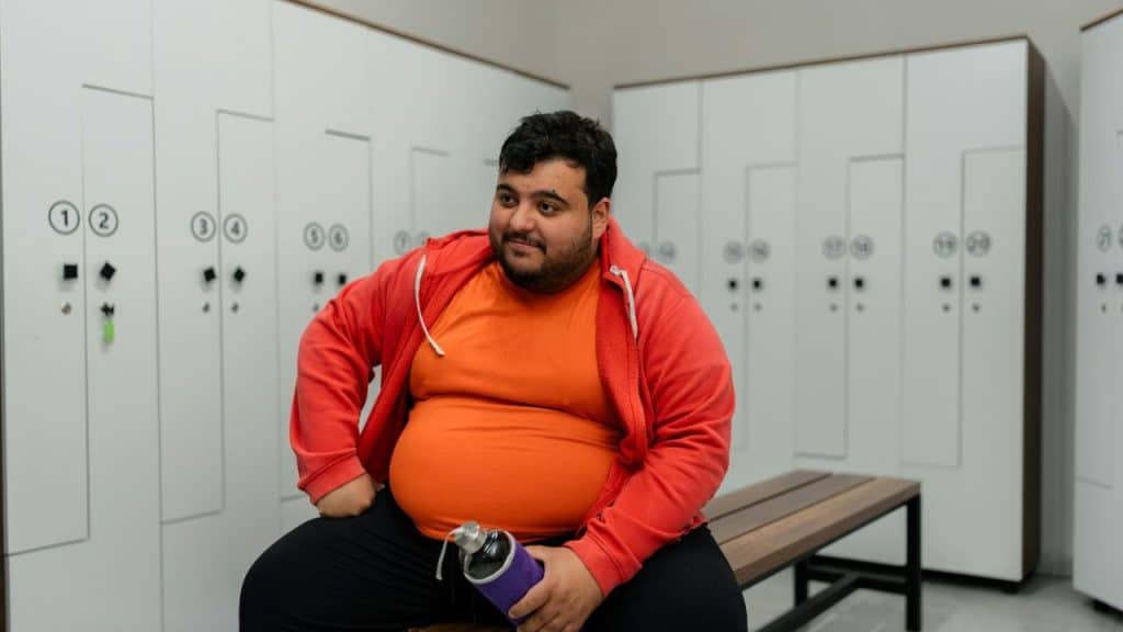 A large man sits on a bench in a locker room, holding a water bottle.