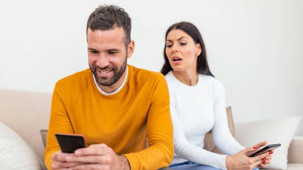 A man smiles at his phone while a woman behind him looks annoyed at her phone.