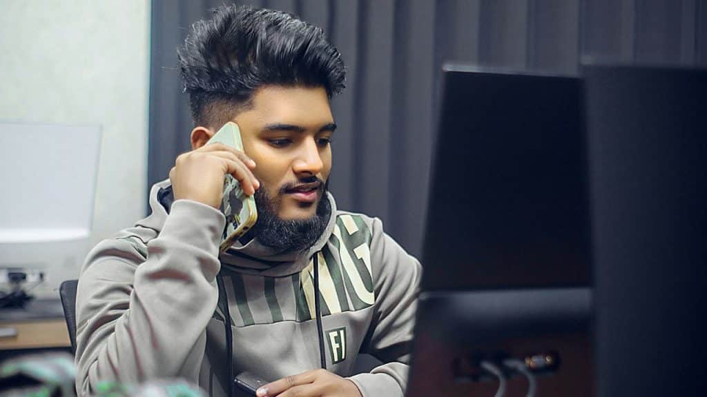 A man with dark hair and a beard speaks on a phone, looking at a computer screen.