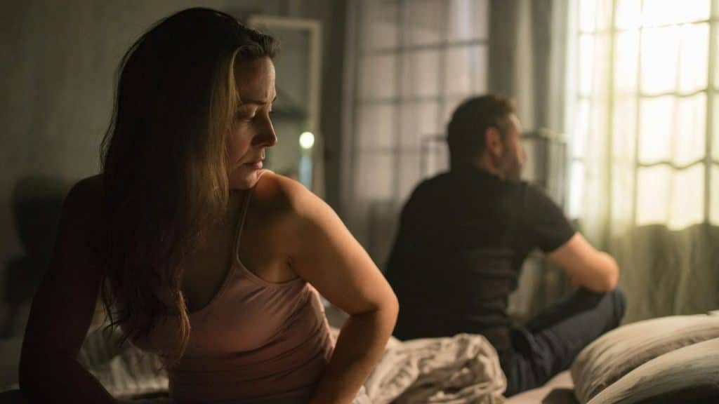 A woman in a tank top sits on a bed, looking away from a man in the background.