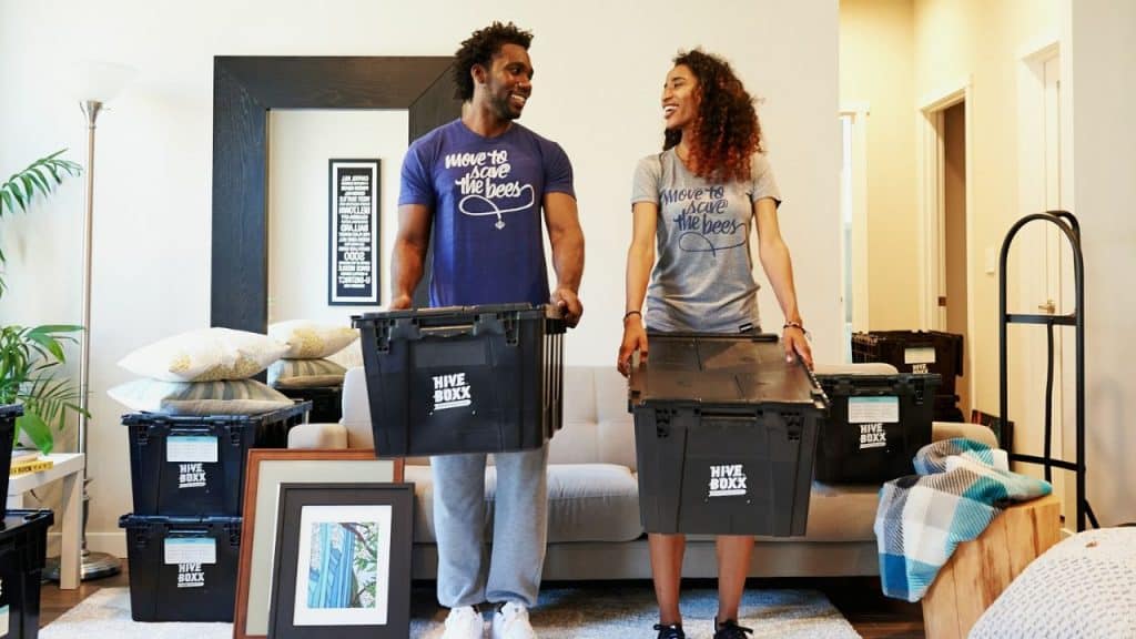 A smiling couple carries moving boxes in a living room filled with packed belongings.