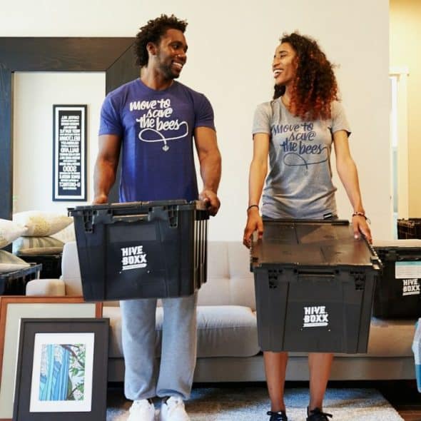 A smiling couple carries moving boxes in a living room filled with packed belongings.