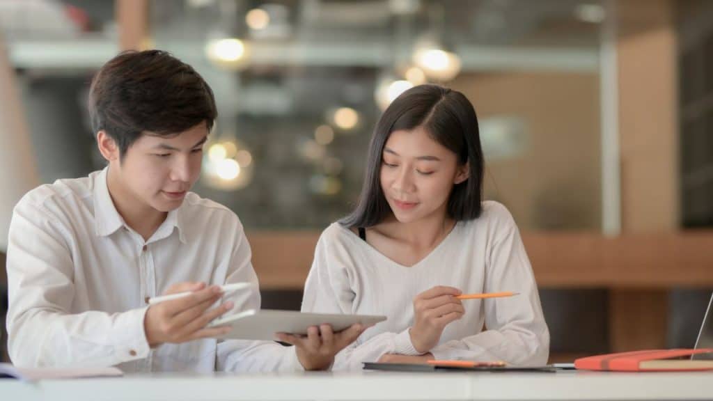 A man and a woman in white shirts look at a tablet together, with a laptop nearby.