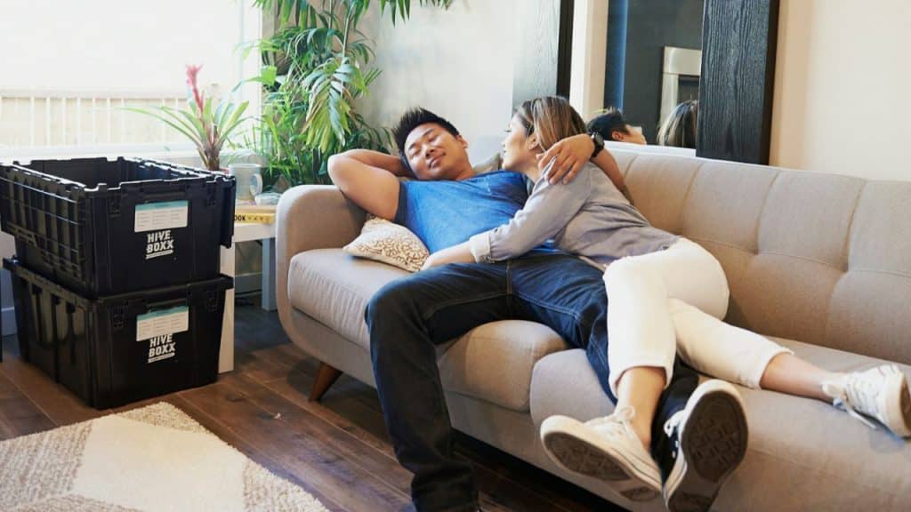 A man lies on a couch with his arm behind his head, while a woman lies next to him with her arm around him.