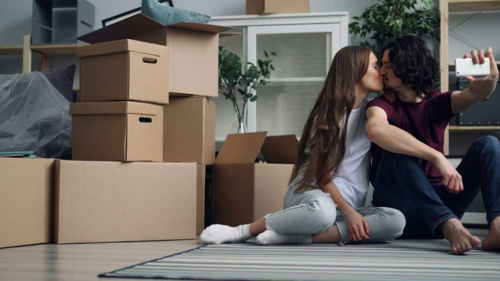 A couple kisses while taking a selfie on the floor surrounded by moving boxes.