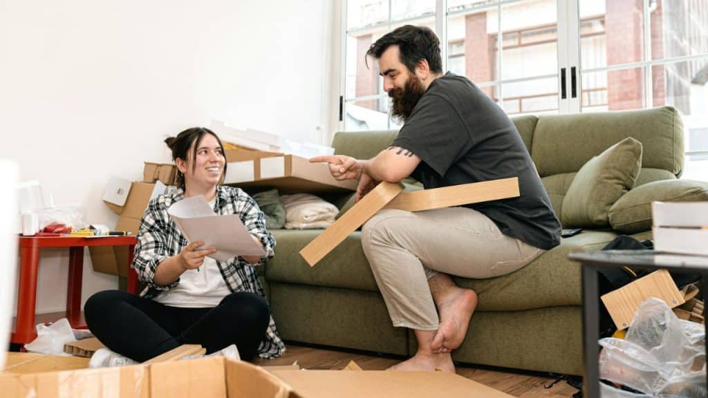 A bearded man and a woman are assembling furniture on the floor in a room with boxes and a sofa.