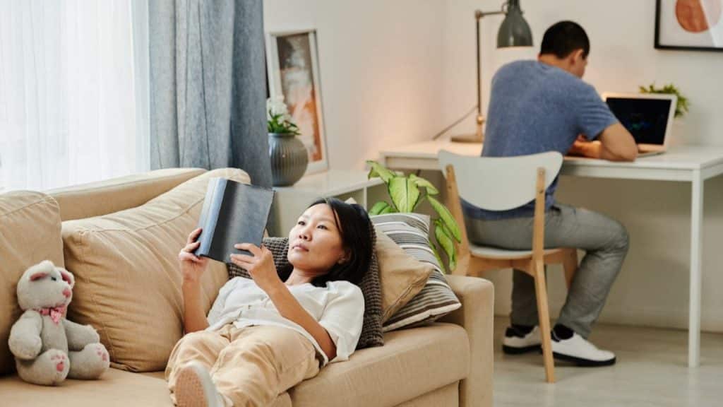 A woman reads on a sofa while a man works at a desk in the background.
