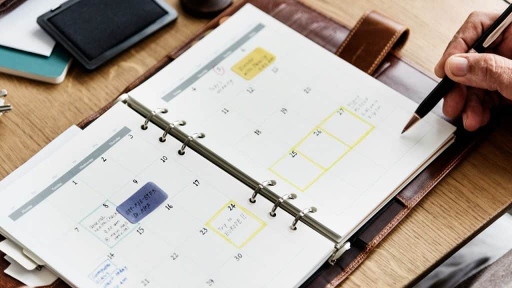 A hand writes on an open planner showing a calendar with highlighted dates.
