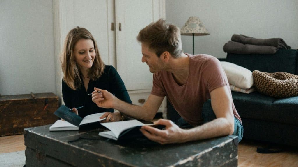A smiling couple sits on the floor, looking at notebooks and discussing something.